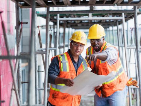 Two multi-ethnic men working at a construction side wearing hard hats, safety glasses and reflective vests, looking at plans. They are inside the structure being built. The taller one is a mid adult African-American man in his 30s. The other one, holding the plans, is a young Hispanic man in his 20s.