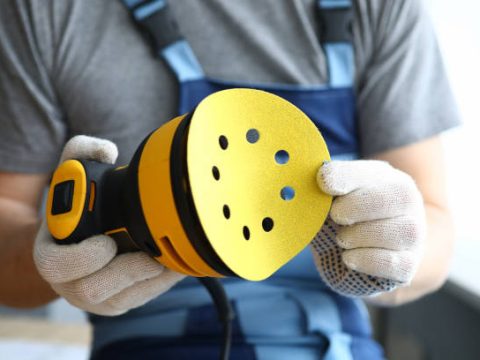 A construction tool for polishing and grinding wood surfaces. A man in gloves holds a spare sole for a grinding machine